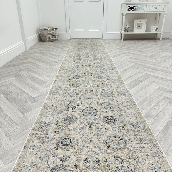 The Analise 7604-580 Runner Rug, featuring a long, patterned design in grey and beige, is laid on a light herringbone wood floor in a bright hallway with a white door, basket, and mirrored console table in the background.