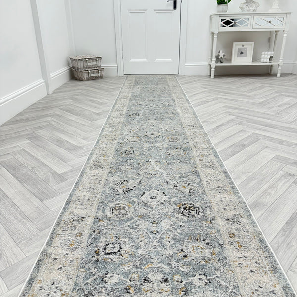The Analise 7604-580 Runner Rug, featuring a long grey pattern, lies on herringbone light wood in a bright hallway with a closed white door, mirrored console table, and wicker basket against the walls.