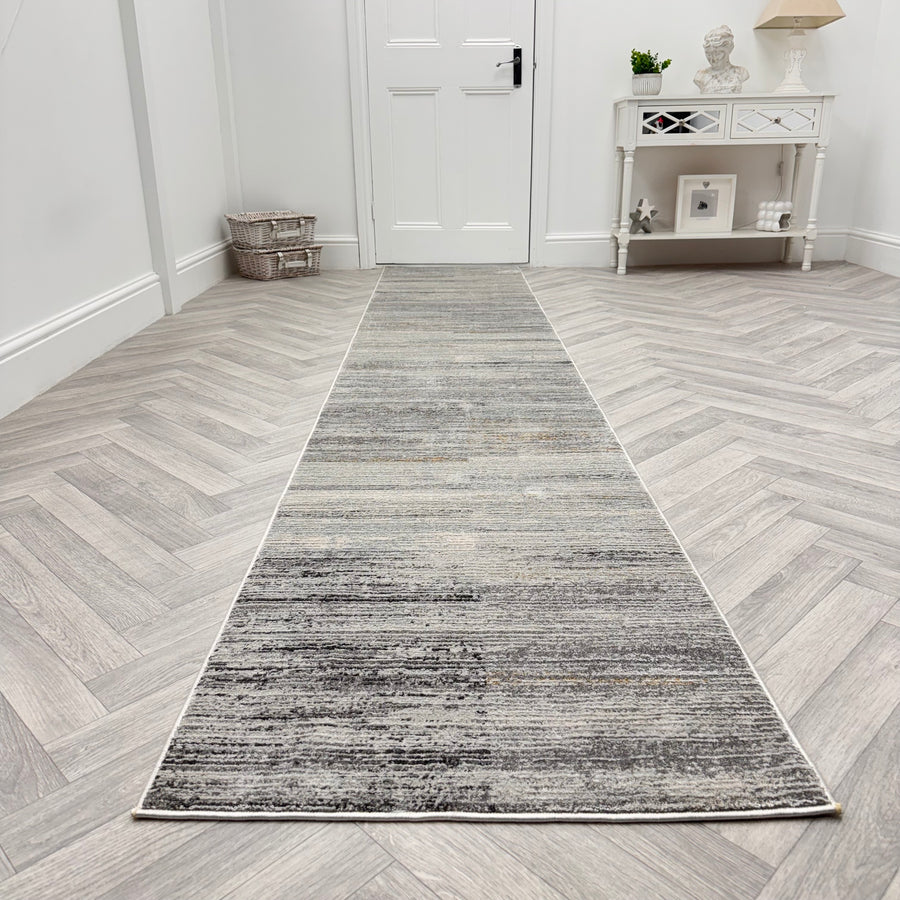 The Analise 7611-989 Runner Rug, featuring a subtle gray pattern, lies on a light herringbone floor in a bright hallway. A white console table with decor and a small basket complements the elegant rug near the back wall.