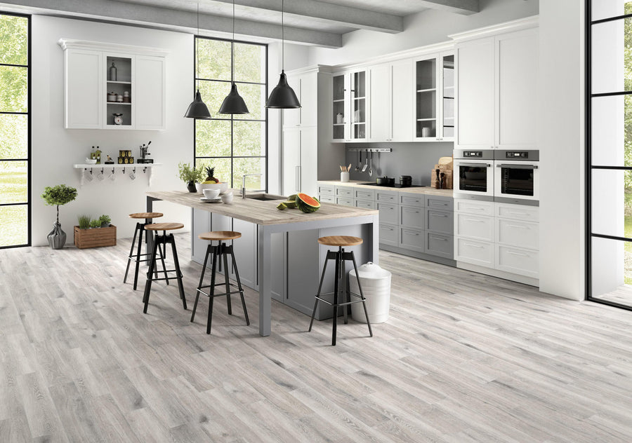 Modern kitchen with white cabinets, large windows, a gray island with wooden stools, pendant lights, and light wood flooring. A watermelon sits on the island, and trees are visible outside.