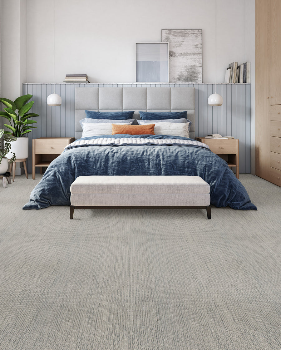 A modern bedroom features a large bed with blue bedding, a gray headboard, and an orange accent pillow. Two wooden nightstands with lamps flank the bed. A bench sits at the foot, and a potted plant is on the left.