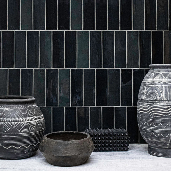 Four black clay pots with white geometric patterns sit on a light surface in front of a wall covered in glossy, rectangular black tiles arranged vertically.