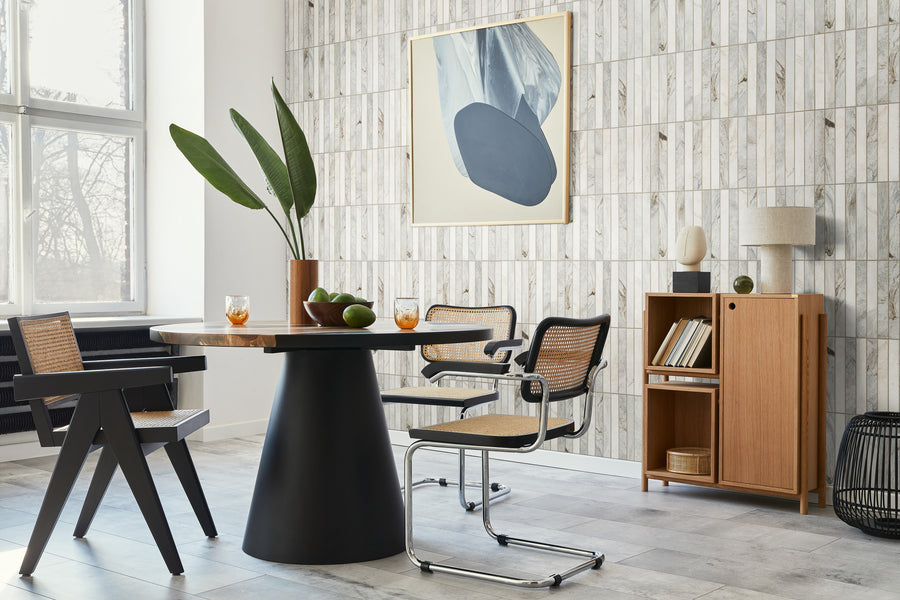 A modern dining room with a round black table, four chairs with woven seats, a plant centerpiece, drinks, and limes. Light wood and cane furniture and abstract art decorate the space. Large window and natural light.