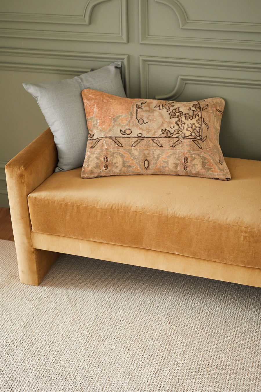 A mustard yellow velvet bench with a beige patterned pillow and a gray pillow, set against a sage green paneled wall and placed on a light textured rug.