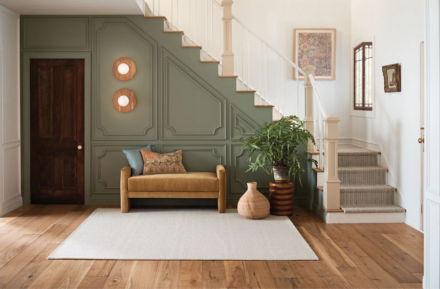 A cozy entryway with a wooden bench and pillows sits against a sage-green paneled wall under a staircase. Decor includes two round wall hangings, a potted plant, a woven vase, and a light-colored area rug on wood flooring.