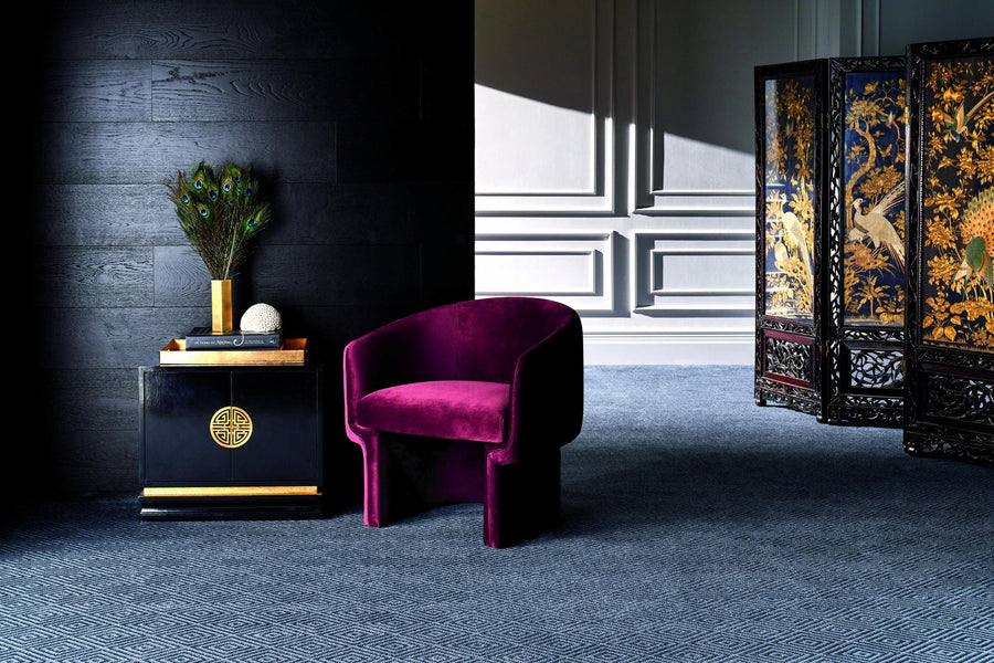 A modern room with a deep purple velvet chair beside a black side table holding peacock feathers in a vase, a book, and a decorative sphere. An ornate gold and black folding screen stands in the background.