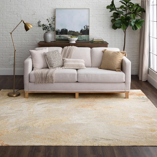 A bright, modern living room with a light gray sofa, neutral throw pillows and blanket, a gold floor lamp, a sideboard with art and decor, a potted plant, and a large, soft patterned rug on dark wood floors.