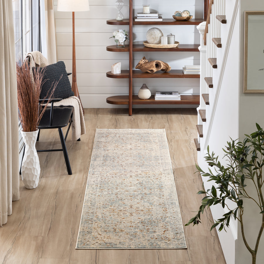 A hallway with light wood floors, a beige patterned runner rug, a black chair with a pillow, a white vase with tall brown branches, a floor lamp, and a wooden shelf with decorative items near a white staircase.