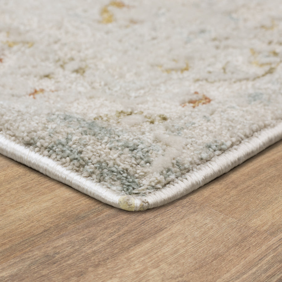 Close-up of the corner of a light-colored, textured area rug with subtle patterns, placed on a wood floor. The rug has a finished edge and sits flat against the smooth surface.