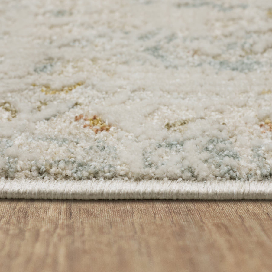 Close-up side view of a light-colored, plush area rug with a subtle pattern, resting on a wooden floor. The texture of the rug and its neatly finished edge are clearly visible.