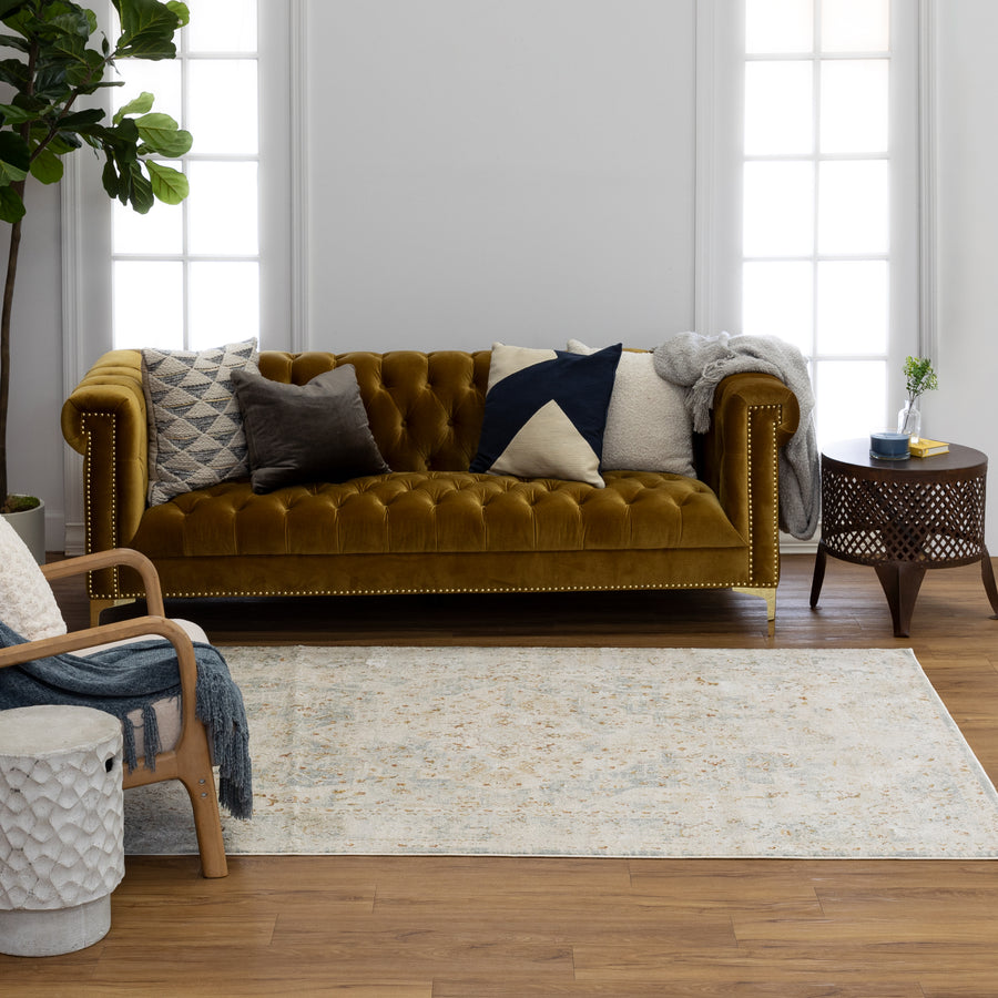 A living room with a mustard yellow tufted sofa decorated with assorted pillows, a wooden side table, a potted plant, an armchair with a blue throw, and a light patterned rug on wooden floor.