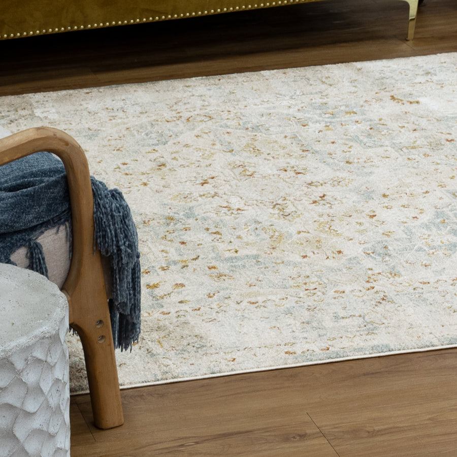 A textured beige area rug with subtle patterns lies on a wood floor, next to a cushioned wooden chair draped with a folded blue blanket and a round, light gray ottoman.