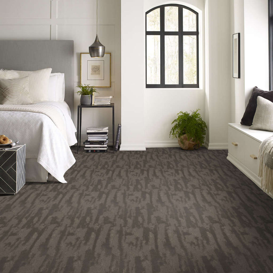 A modern bedroom with a gray patterned carpet, white bedding, a nightstand with books and a lamp, a potted plant, and a window seat under a black-framed window letting in natural light.