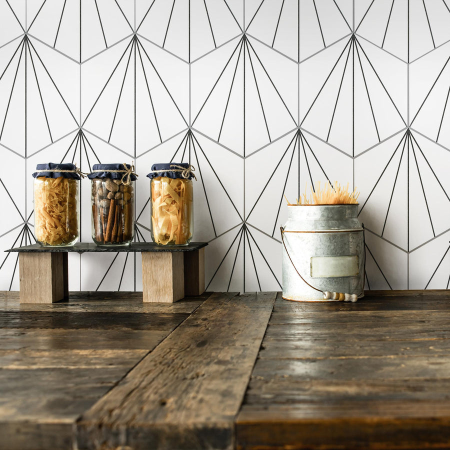 Three glass jars filled with dried ingredients sit on wooden blocks next to a metal container holding pasta, all on a rustic wooden counter with a geometric patterned white tile wall behind them.