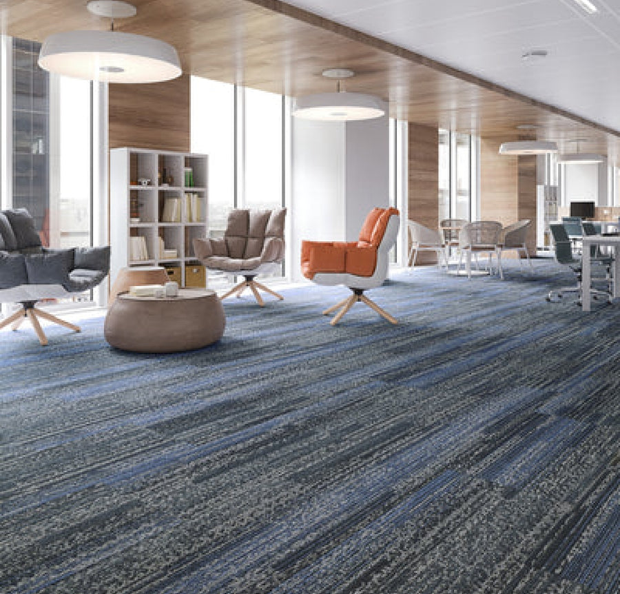 A modern office lounge features floor-to-ceiling windows, armchairs, a round ottoman, bookshelf, round tables, and Commercial Modular Carpet Tiles in a blue pattern. Abundant natural light creates a bright and welcoming environment.