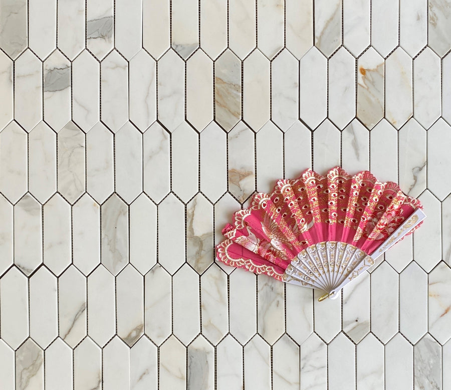 A decorative pink and gold hand fan with lace trim is displayed on a white and gray marble tile surface with a geometric pattern.