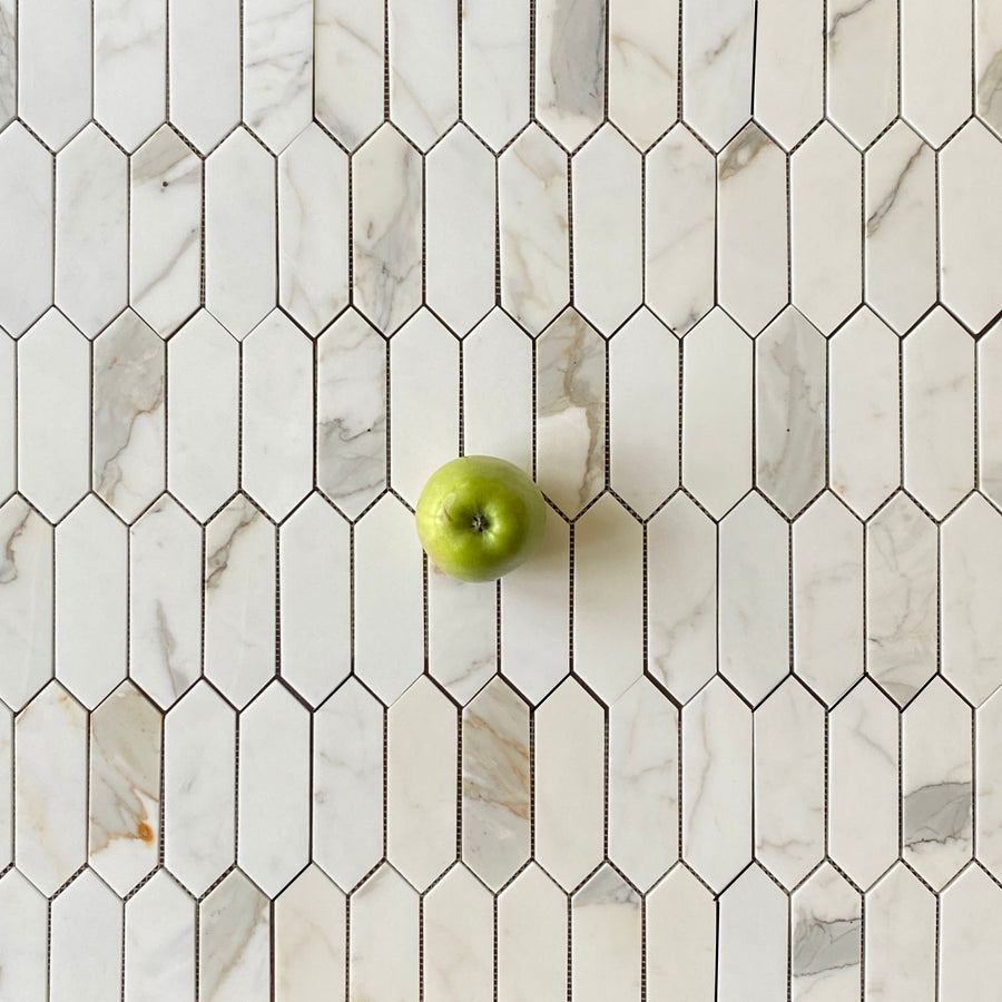 A single green apple is placed on a white, elongated hexagon-patterned marble tile surface with grey and tan veining.