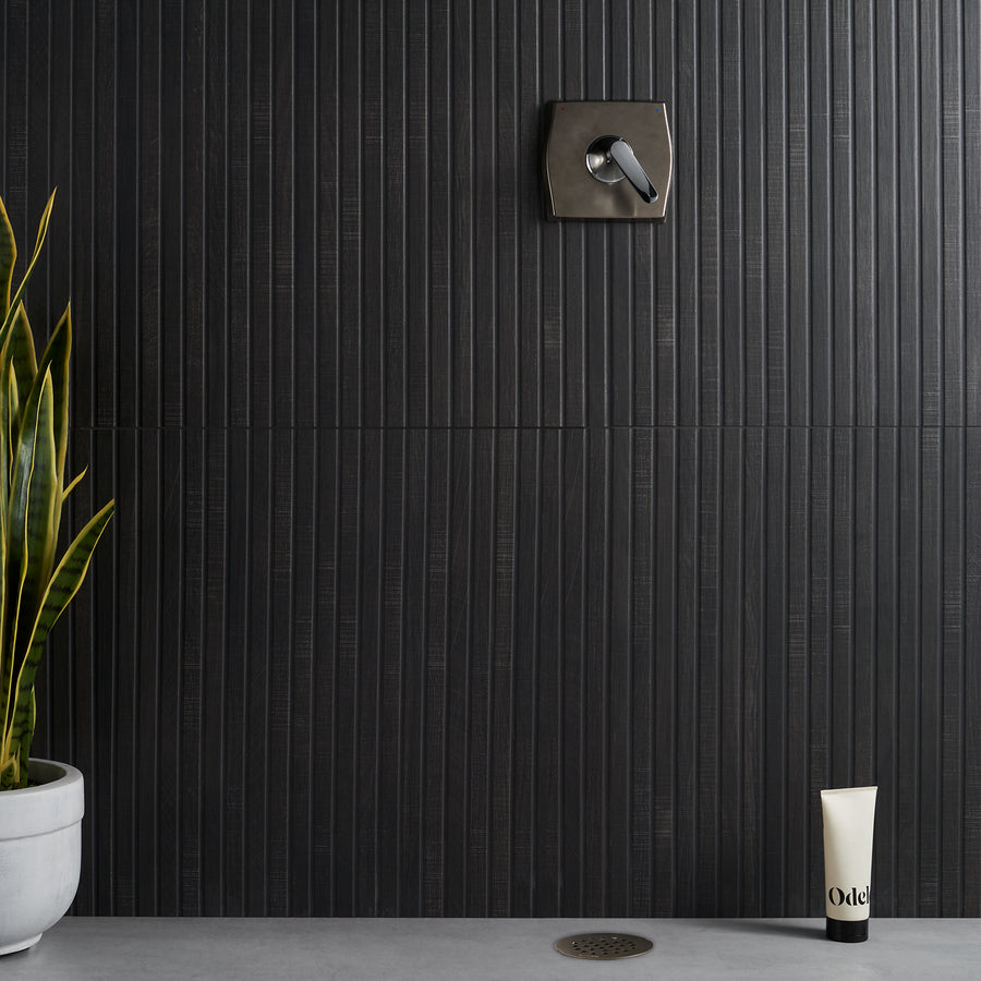A modern shower with a dark vertical tile wall, a metal control handle, a potted snake plant on the left, a tube of Odé product on the floor, and a round drain in a concrete floor.