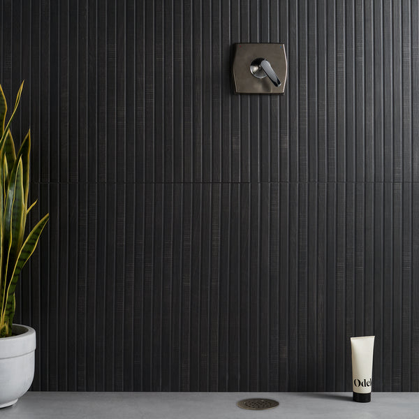 A modern shower with a dark vertical tile wall, a metal control handle, a potted snake plant on the left, a tube of Odé product on the floor, and a round drain in a concrete floor.