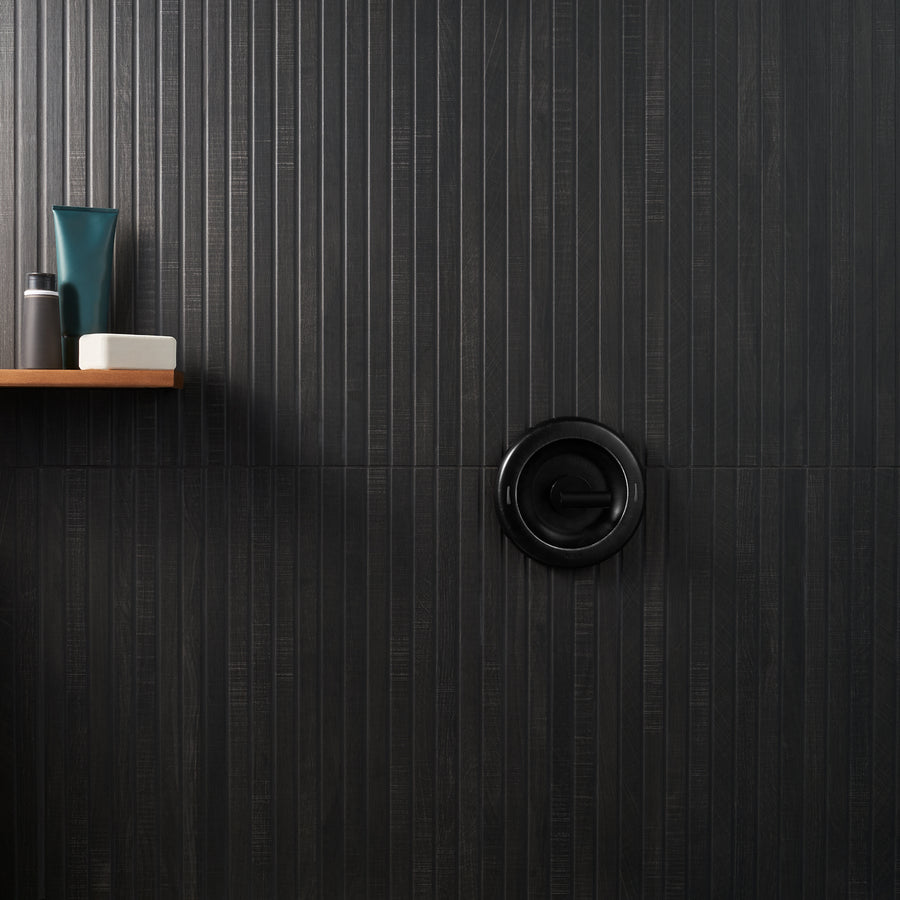 A modern shower with a matte black control handle on a dark, vertically grooved tile wall. On a small wooden shelf to the left are bottles and a bar of soap.