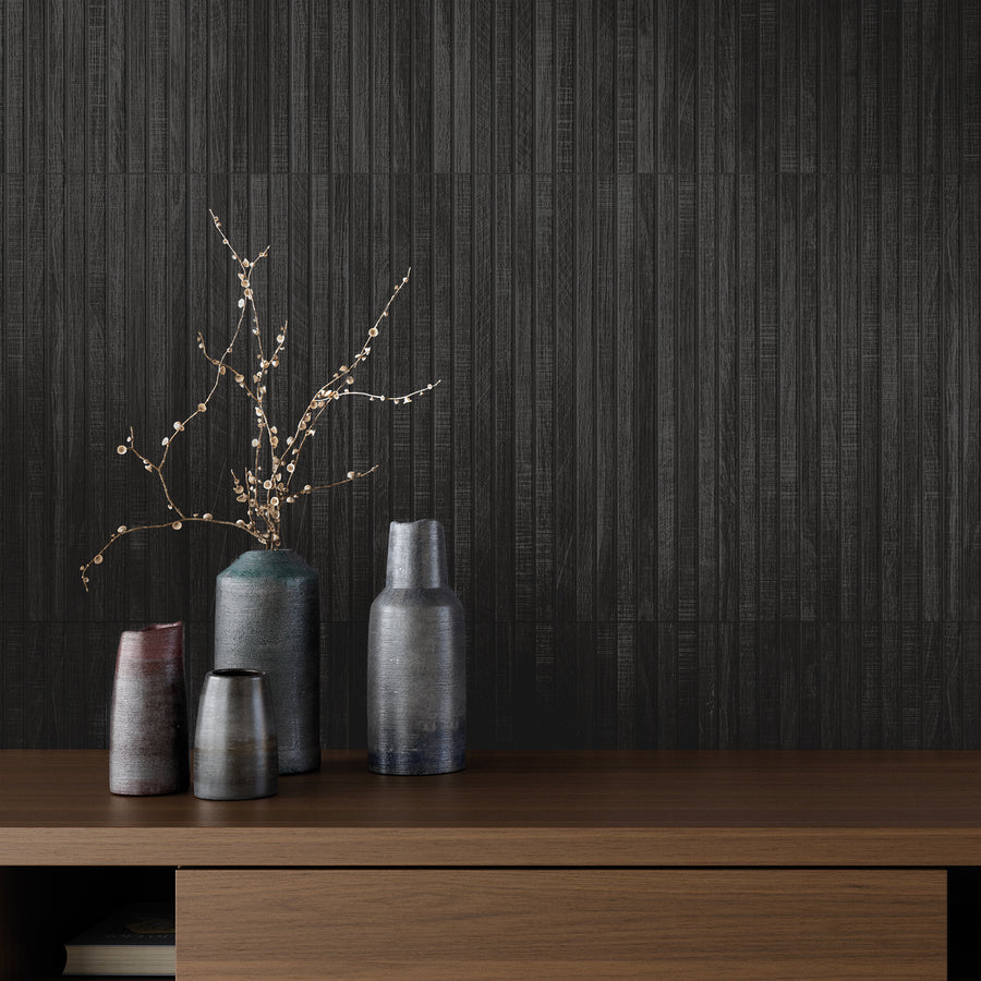 Four ceramic vases in varying sizes and colors sit on a wooden surface; one vase holds delicate branches with small white buds, set against a dark, textured vertical-panel wall.