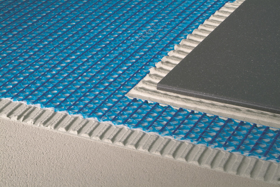 A close-up of Blanke Permat blue uncoupling membrane installed over a gray subfloor with ridged thinset, and a black ceramic tile partially set on top during installation.