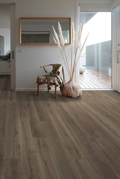 A modern, minimal living space with wood-look flooring, a wooden chair draped with a blanket, a large mirror on the wall, and a vase of pampas grass near a glass door opening to a sunlit deck.