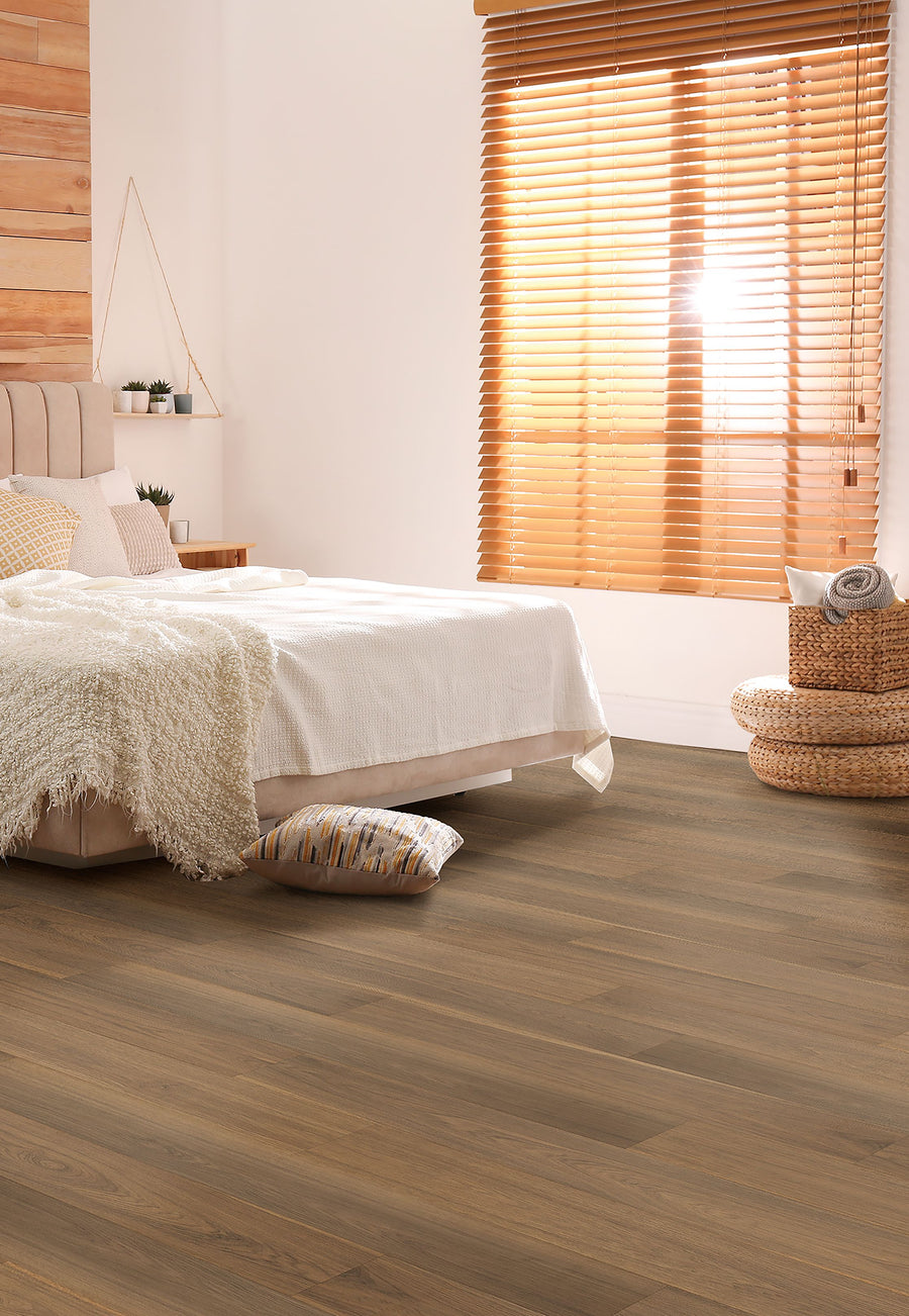 A cozy bedroom with a bed covered in a white blanket, neutral pillows, a soft beige throw, woven baskets, and large windows with wooden blinds letting in warm sunlight. The floor is finished with light wood planks.
