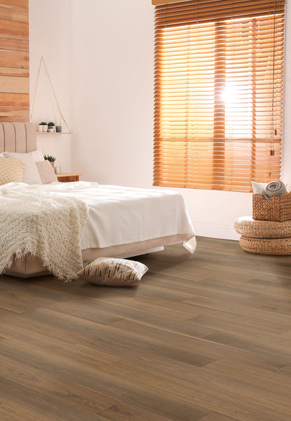 A cozy bedroom with a bed covered in a white blanket, neutral pillows, a soft beige throw, woven baskets, and large windows with wooden blinds letting in warm sunlight. The floor is finished with light wood planks.