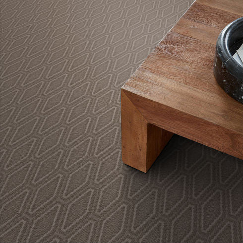 A geometric-patterned, taupe carpet next to the corner of a wooden coffee table with a round black bowl on top.