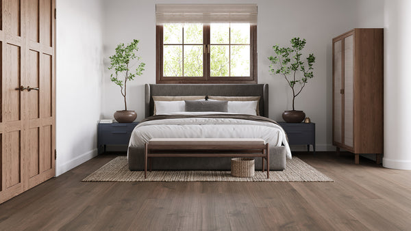 A modern bedroom with a large bed, gray headboard, blue nightstands, potted plants, wooden wardrobe, and bench. There’s a window above the bed, neutral decor, and natural light brightening the room.