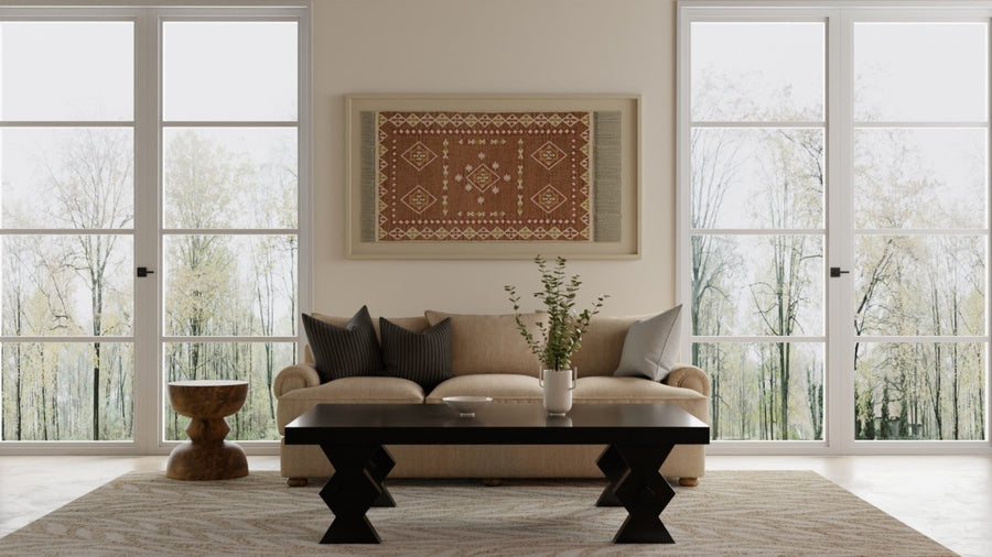 A beige sofa with gray and beige pillows sits in front of large windows in a bright living room. A black coffee table, small wooden side table, and a decorative rug hanging on the wall complete the modern decor.
