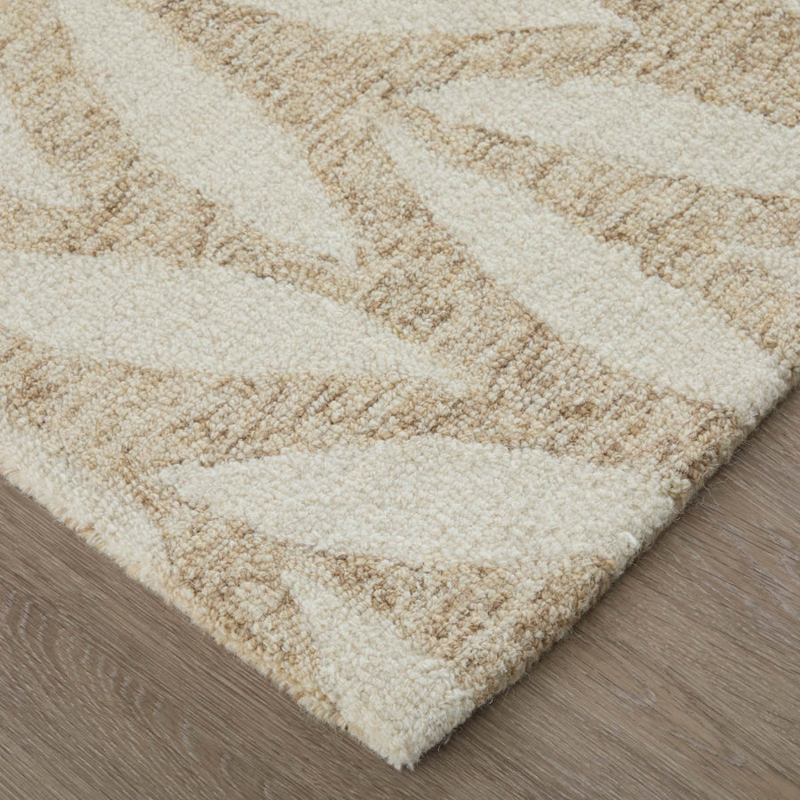 A close-up of a beige and light brown patterned rug with a raised textured design, placed on a light wood floor. The rug features abstract shapes and a soft, plush appearance.