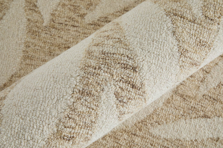 Close-up of a textured, beige and cream area rug with a subtle, abstract leaf or vine pattern; the rug is partially rolled, showing its plush, woven surface.