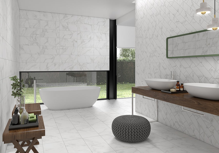 Modern bathroom with white marble walls and floor, freestanding bathtub by a large window, double sinks on a wood vanity, round gray pouf, green-framed mirror, and decorative plants and toiletries.