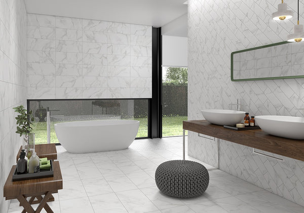 Modern bathroom with white marble walls and floor, freestanding bathtub by a large window, double sinks on a wood vanity, round gray pouf, green-framed mirror, and decorative plants and toiletries.