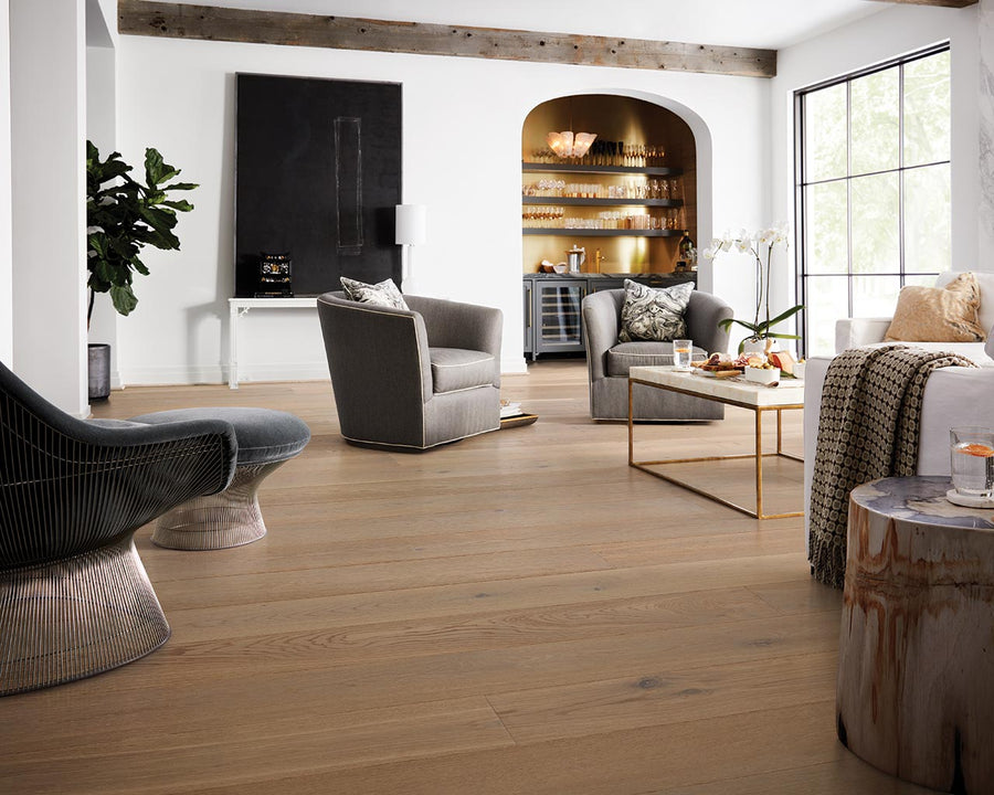 A modern living room with light wood floors, gray and cream furniture, a large window, and built-in shelves with glassware. A potted plant, abstract wall art, and gold accents add style to the bright, airy space.