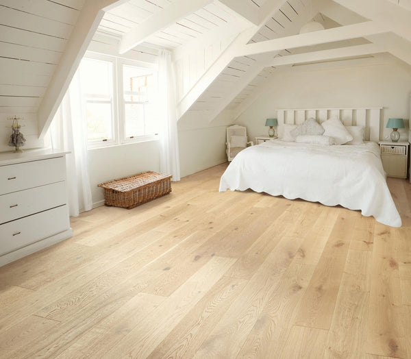 Bright attic bedroom with large windows, white walls and ceiling, light wooden floor, a double bed with white bedding, bedside tables with lamps, a wicker chest, and a dresser.