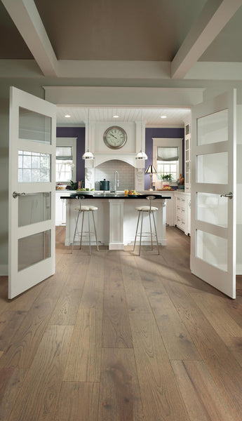 Open double glass doors lead to a modern kitchen with a central island, barstools, pendant lights, a wall clock, and light wood flooring. The kitchen features white cabinetry and neutral tones.