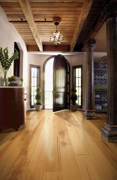Elegant entryway with light wood flooring, an arched dark wooden front door open to the outside, potted plants, a rustic wooden ceiling, exposed beams, and decorative columns. Sunlight streams in, creating a warm atmosphere.
