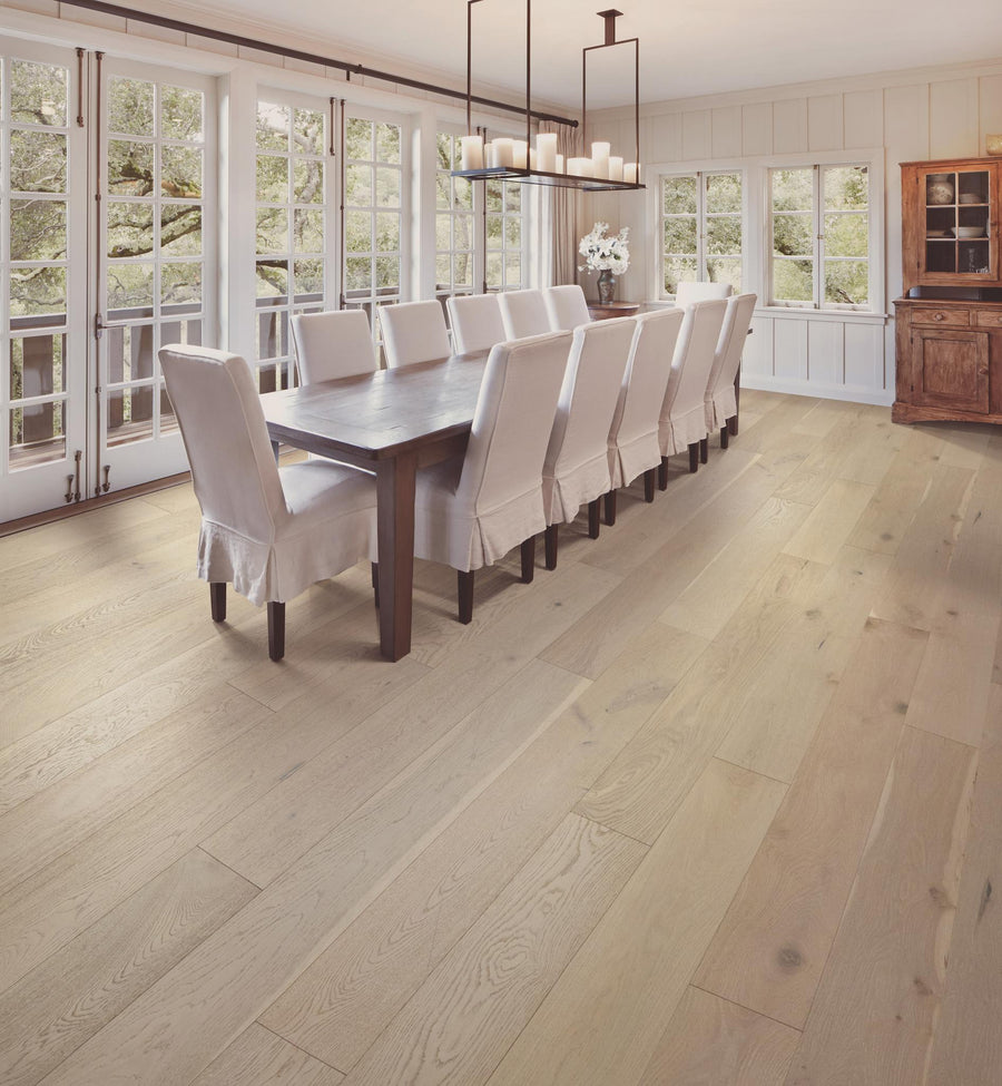 Bright dining room with large windows, a long dark wood table, twelve white slipcovered chairs, a modern black chandelier, and light wood flooring. A wooden cabinet stands in the back corner.