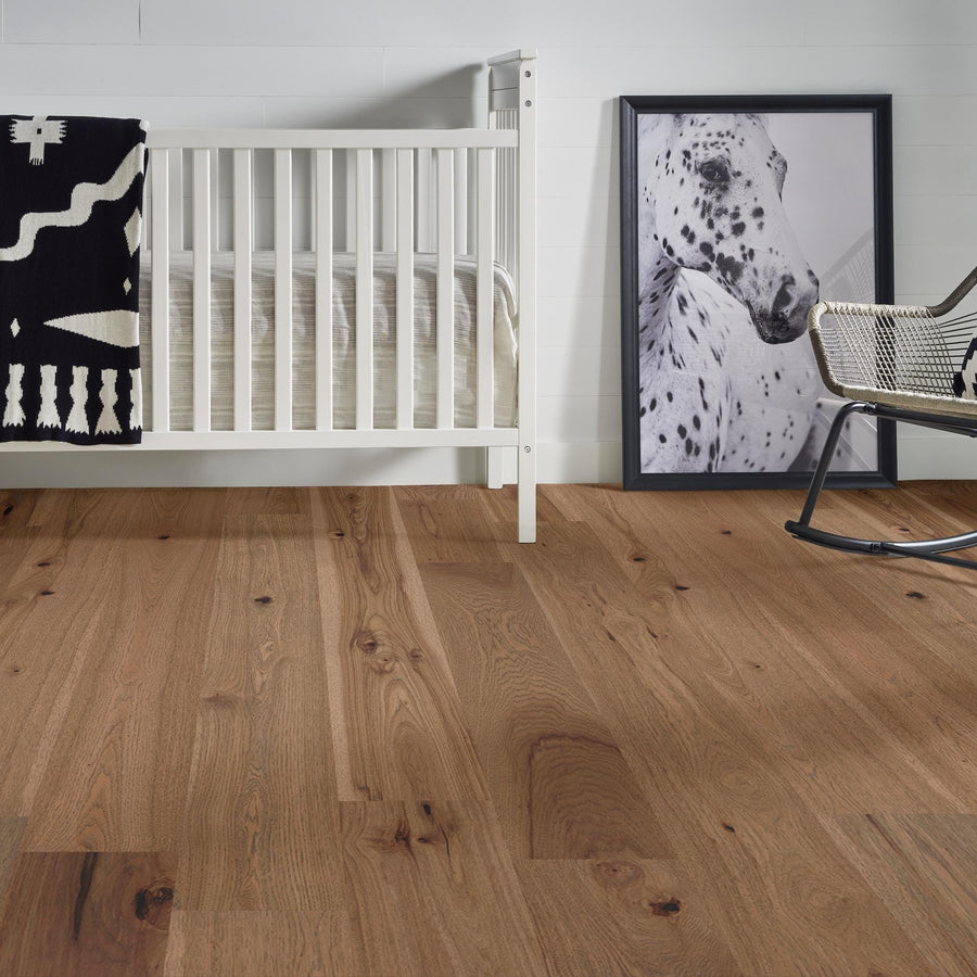 A modern nursery with wooden floors, a white crib with a black-and-white blanket, a framed photo of a spotted dog leaning against the wall, and a metal rocking chair.