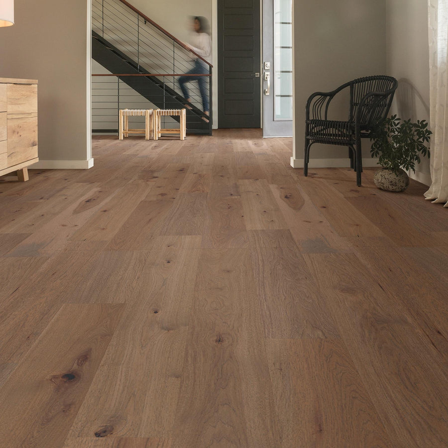 A spacious room with light brown wooden flooring, neutral walls, a black chair, a small plant, and a staircase. A person is walking upstairs, and natural light enters through a window and glass-paneled door.