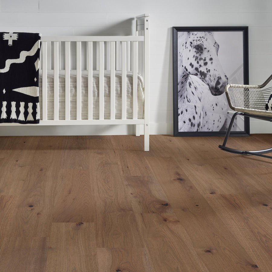 A white crib, a black and white blanket, a framed photo of a spotted horse, and a metal rocking chair sit on a brown wooden floor in a minimalist room with white walls.