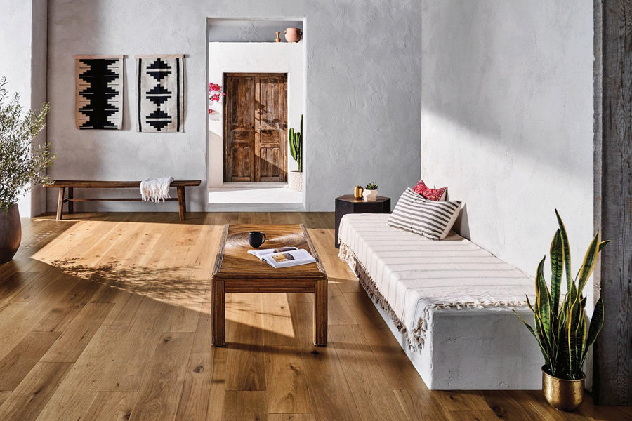 Bright, modern living room with wooden floors, a striped sofa, a wooden coffee table, potted plants, a bench with cushions, geometric wall art, and a rustic wooden door in the background.