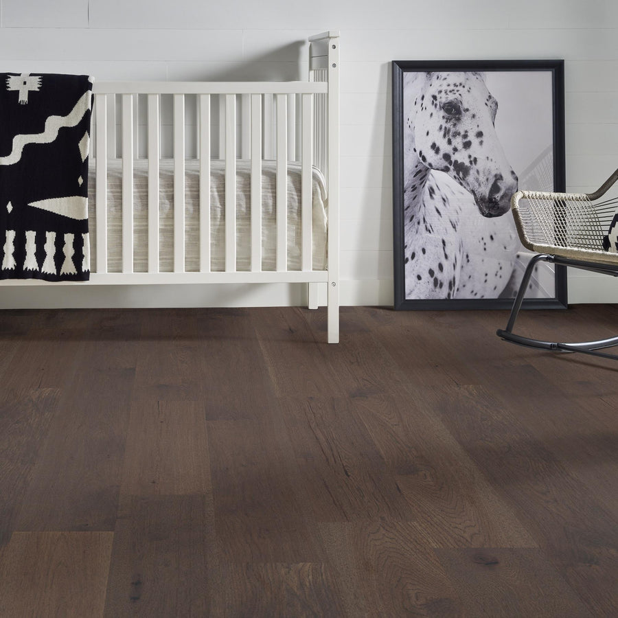 A minimalist nursery with dark wood flooring, a white crib with a black and white patterned blanket, a framed photo of a spotted horse resting against the wall, and a modern metal chair.