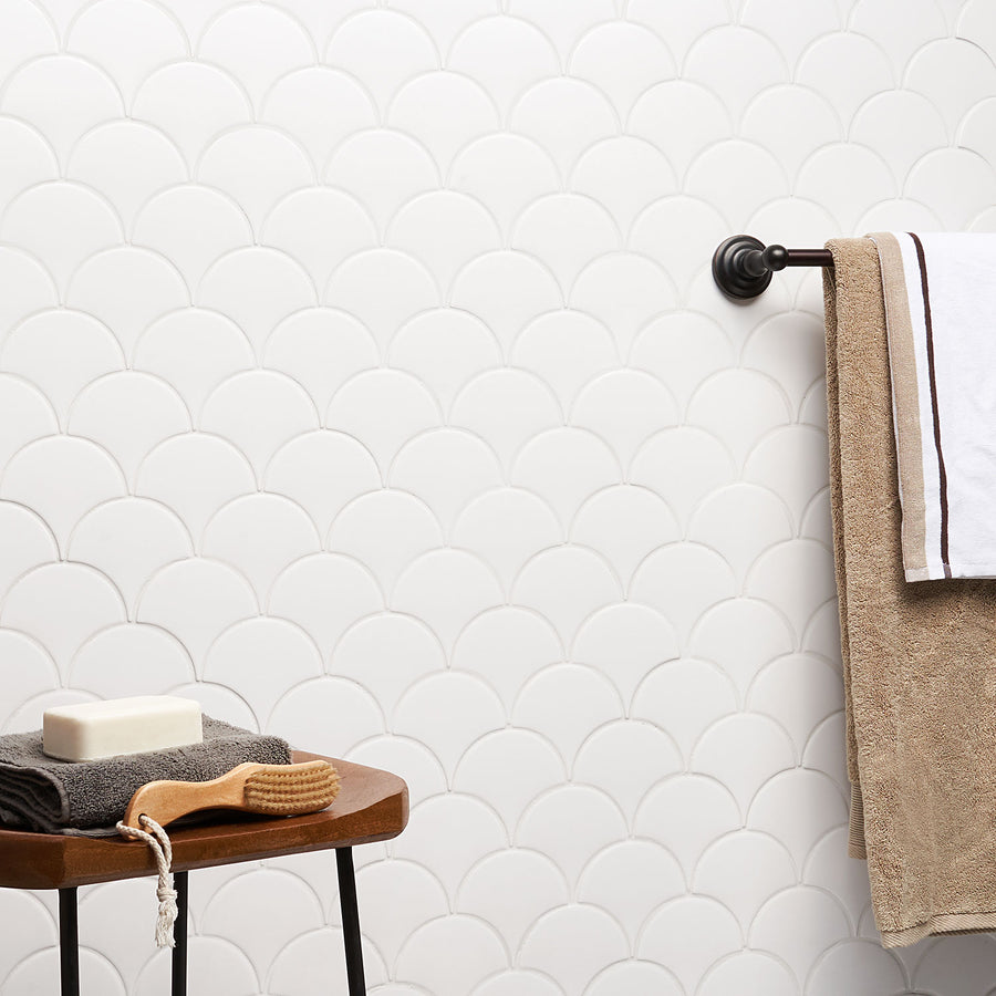 A wooden stool with a folded gray towel, a bar of soap, and a bath brush is next to a wall with white scalloped tiles. A towel rack holds two towels, one tan and one white with brown trim.