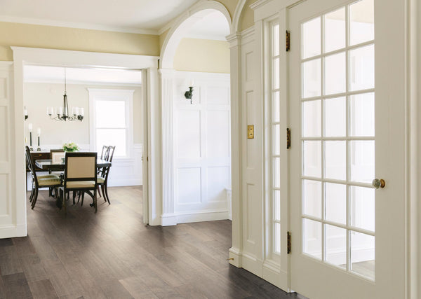 Bright entryway with glass-paneled door and white trim opens to a dining room with a dark wood table, six chairs, candle holders, and windows letting in natural light. The floors are dark wood throughout.