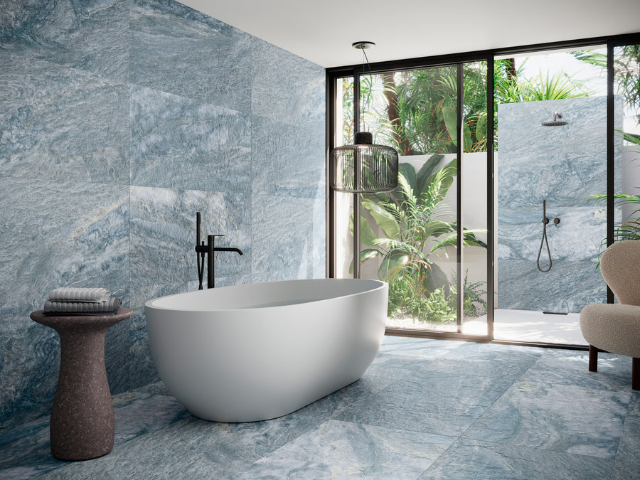 Modern bathroom with a white freestanding bathtub, black fixtures, blue marble-patterned walls and floor, a small side table with towels, and large glass doors opening to an outdoor shower with lush greenery.
