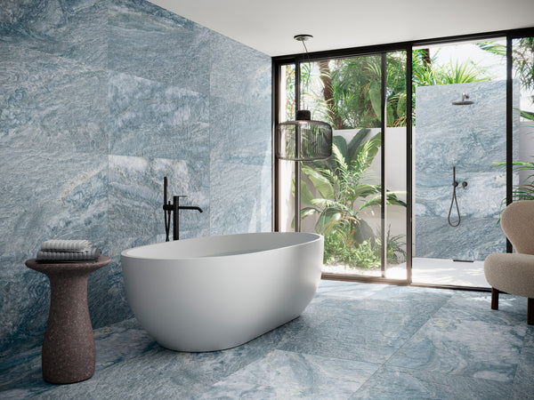 Modern bathroom with a white freestanding bathtub, black fixtures, blue marble-patterned walls and floor, a small side table with towels, and large glass doors opening to an outdoor shower with lush greenery.